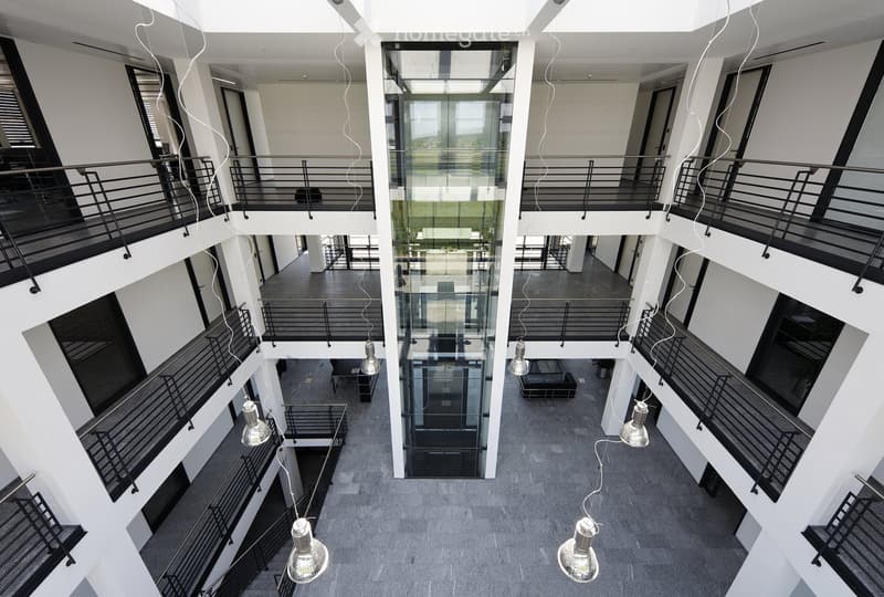 Modern multi-story building interior with open-plan design, glass walls, black metal railings, and hanging pendant lights.