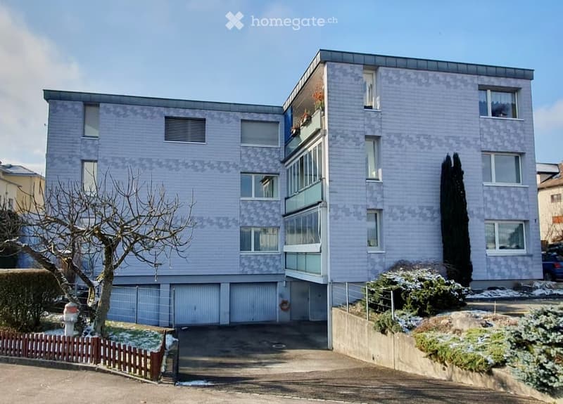 Modern multi-story building with light blue facade, large windows, and a garage entrance at the bottom.