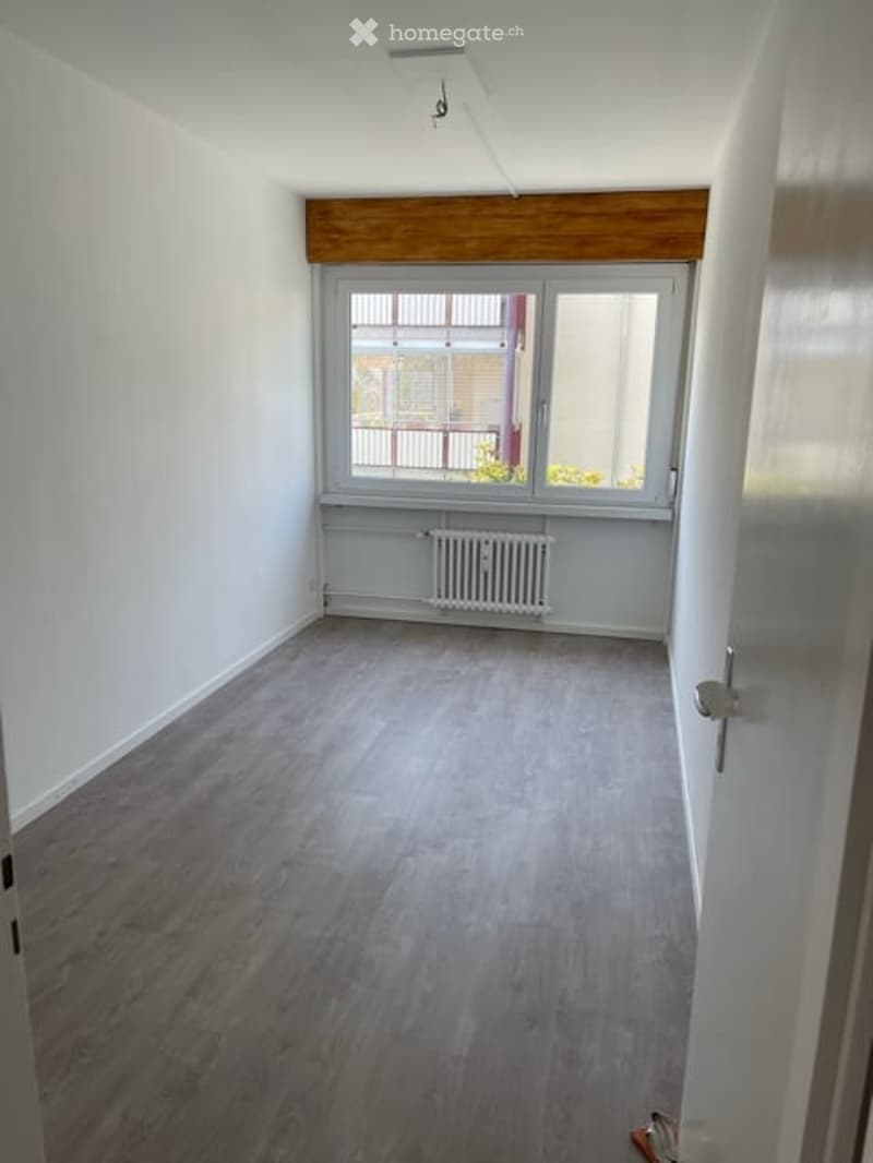 Empty room with light grey wooden flooring, white walls, and a large window with a radiator below. The ceiling has a wooden beam detail.