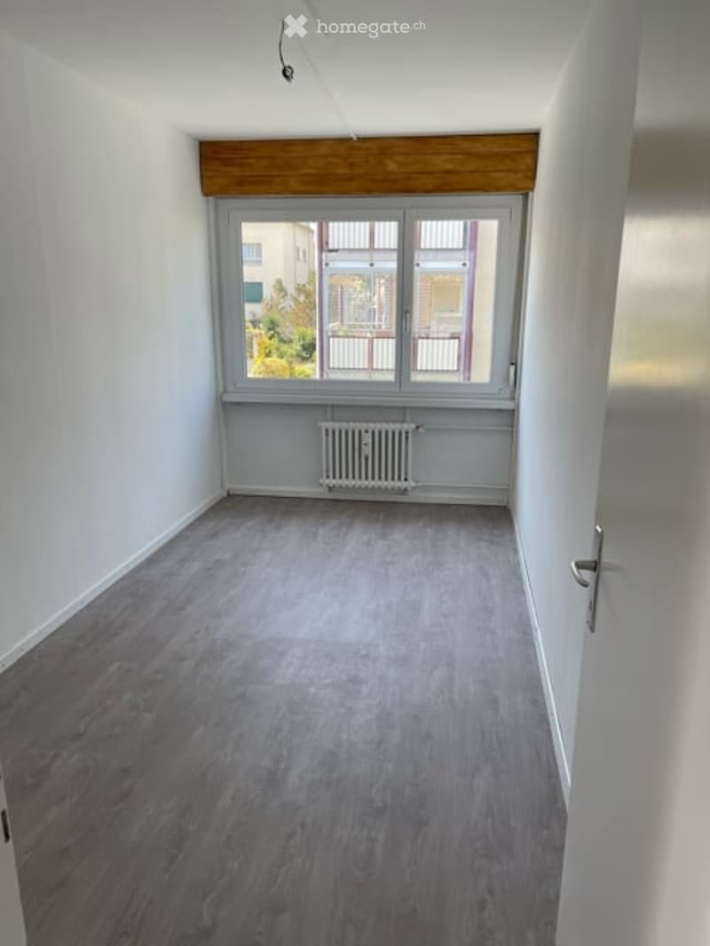 Empty room with light grey wooden flooring, white walls, and a large window with multiple panes. A radiator is visible below the window, and the room has a simple, clean design with no furniture.