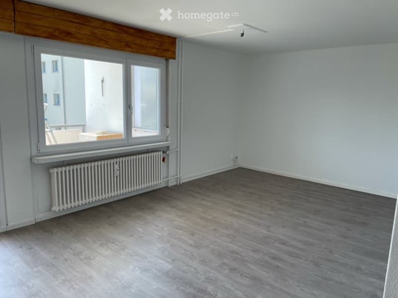 Empty room with light grey walls, dark grey wooden flooring, large window with white frame, radiator below the window, and a wooden ceiling panel.