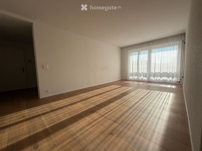 Empty room with wooden flooring, white walls, and large windows with sheer curtains allowing natural light to enter.