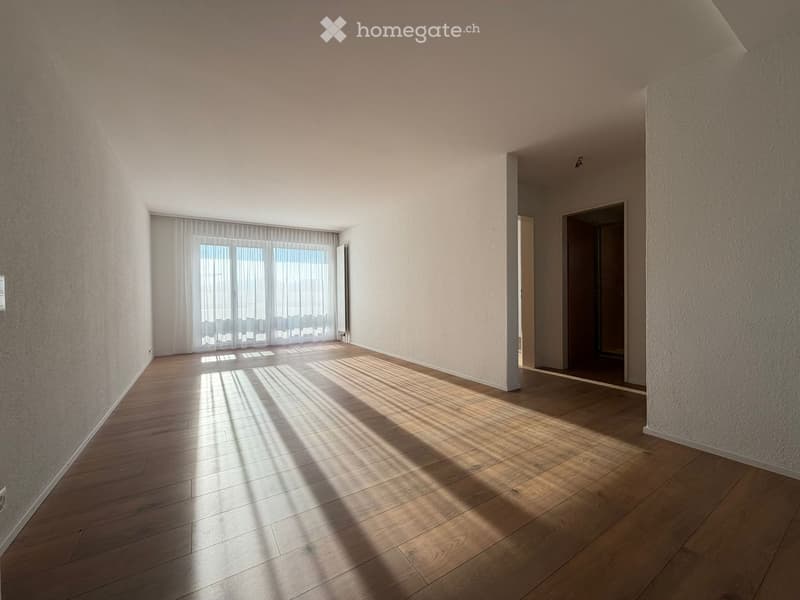 Empty room with wooden flooring, white walls, and large windows with white curtains. Sunlight streams in, casting shadows on the floor. There are two open doorways leading to other spaces.