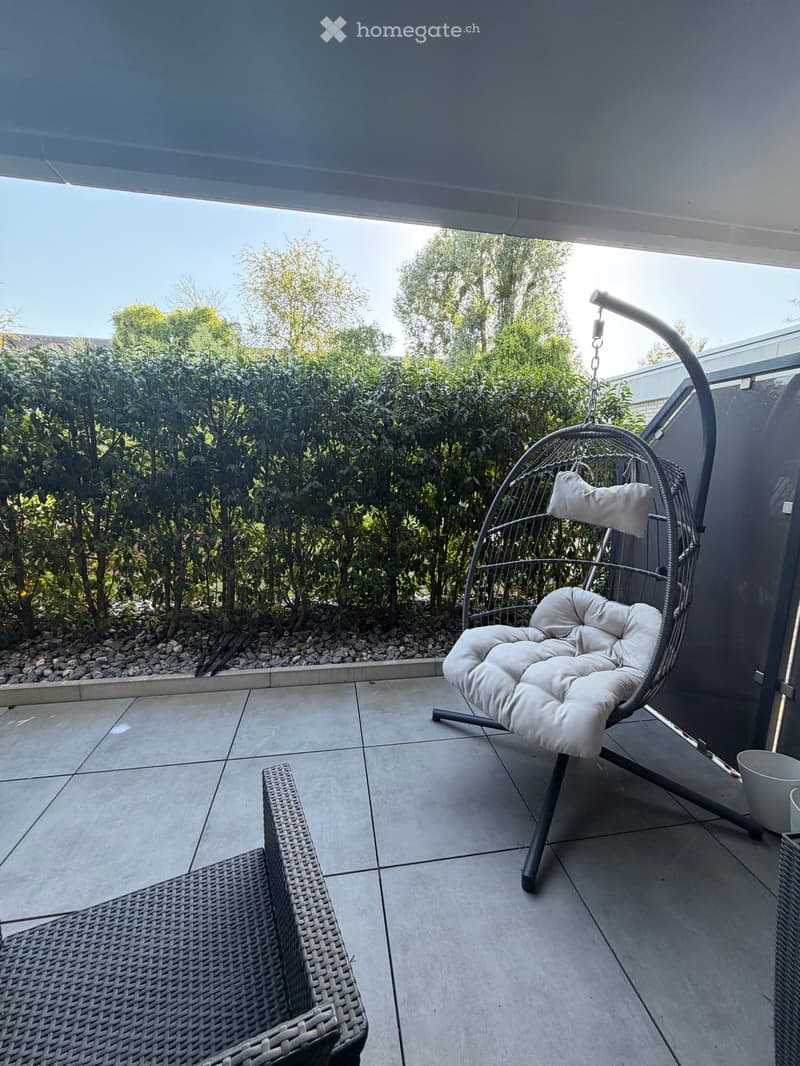 Balcony with tiled floor, wicker chair, hanging egg chair with cushion, and a cup on a small table.