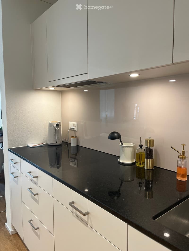 Modern kitchen with white cabinets, black countertop, and various appliances and items including an electric kettle, coffee grinder, and bottles.