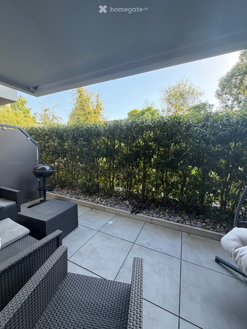 Balcony with wicker furniture, a grill, and a view of greenery.
