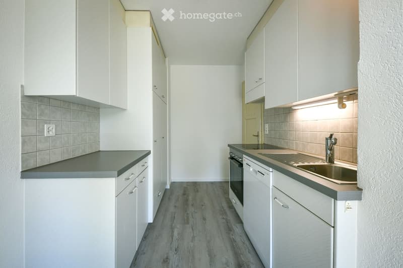Modern kitchen with white cabinets, grey countertops, and a stainless steel sink and oven. The floor is made of wood, and the walls are tiled.