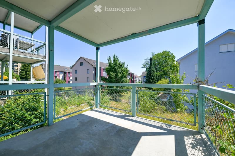Balcony with metal railing, concrete floor, and a view of surrounding buildings and greenery.