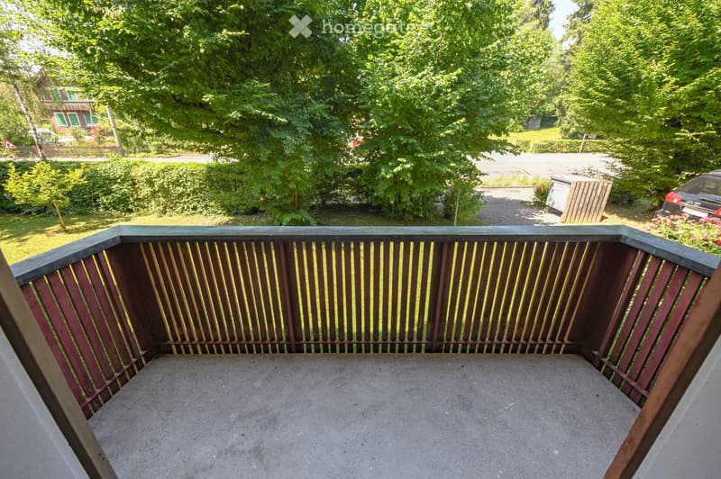 Balcony with wooden railing, concrete floor, overlooking a green garden area with trees and a driveway.