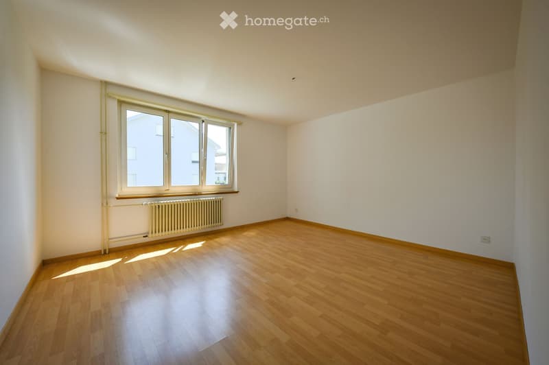 Empty room with wooden flooring, white walls, and a large window with a radiator below it.