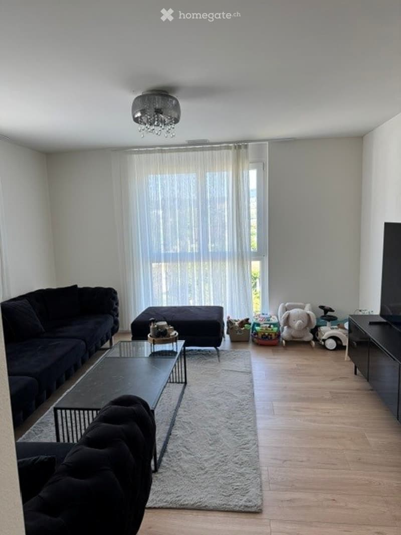 Modern living room with a black sectional sofa, glass coffee table, ottoman, and a TV stand. The room features a large window with sheer curtains, wooden flooring, and a light-colored rug.