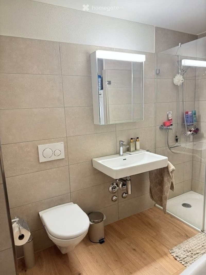 Modern bathroom with a white sink, toilet, mirror, and glass-enclosed shower. Beige tiles, wooden flooring, and a towel hanging on the side.