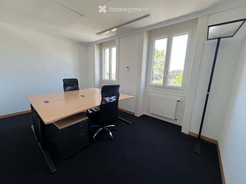 Office space with a large wooden table, two black office chairs, a drawer unit, and large windows letting in natural light.