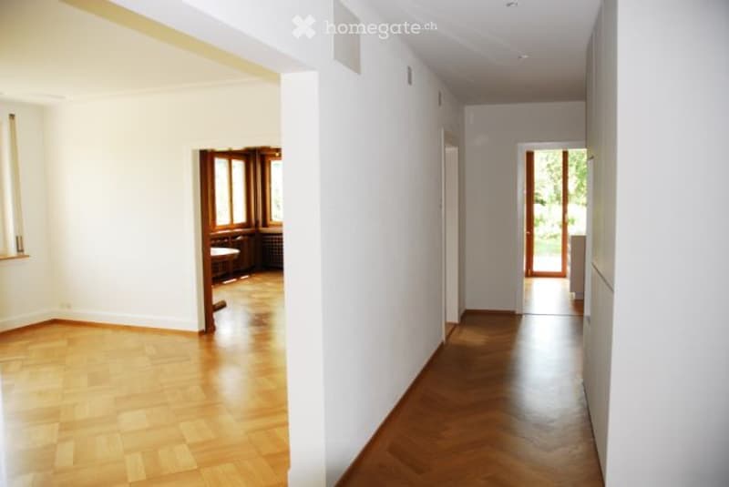 Empty hallway with wooden flooring and white walls, leading to an adjacent room with a small table and chairs.
