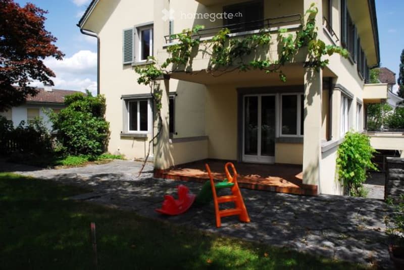 A two-story house with a balcony and a small garden area featuring a slide and a ladder.