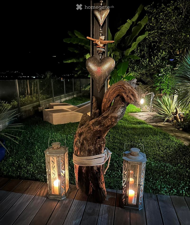 Wooden deck with lanterns, tree trunk with metal hearts, hedges, a glass fence, outdoor seating area