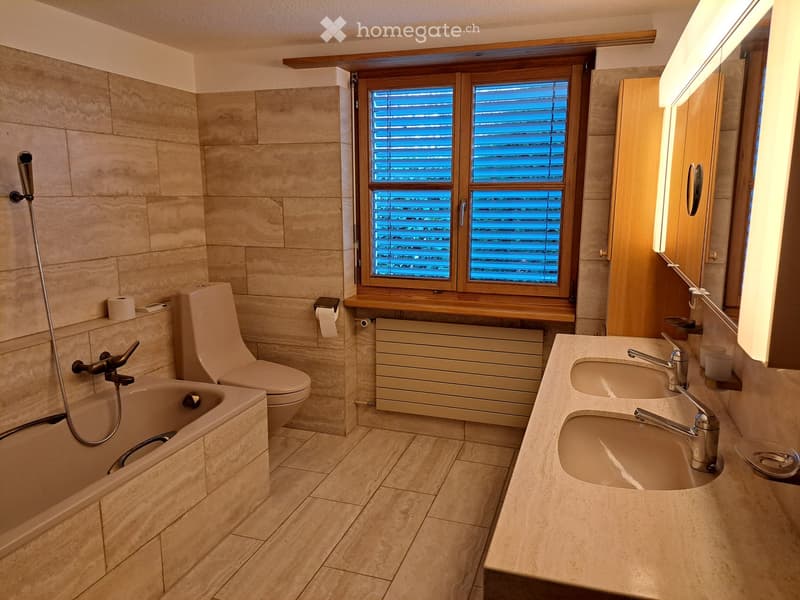 Modern bathroom with beige tiles, a bathtub, toilet, double sink, and a large window with wooden blinds.