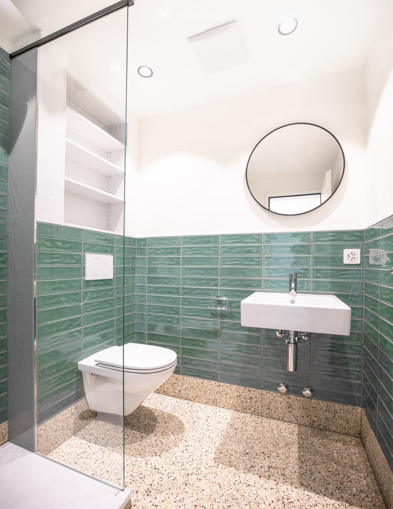Modern bathroom with green tiles, white sink, toilet, and a round mirror above the sink.
