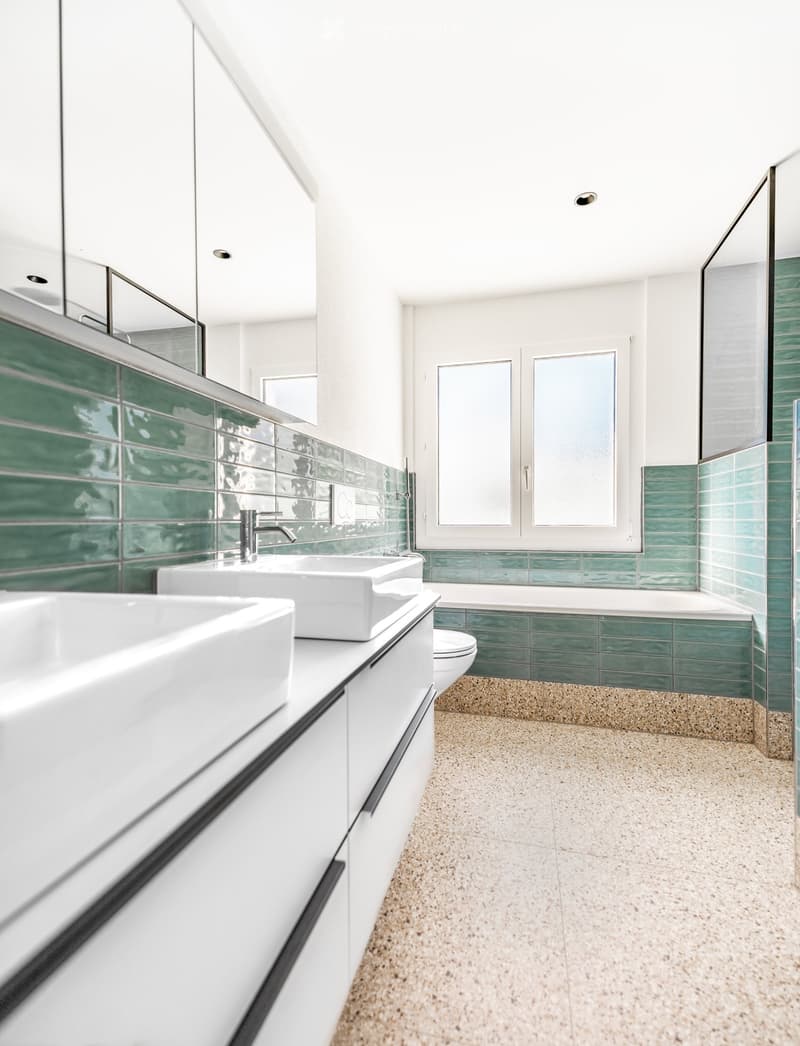 Modern bathroom with white cabinets, double sink, bathtub, and green tiled accents.