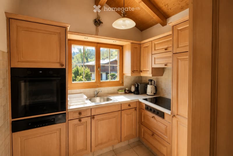 A cozy kitchen with light wooden cabinets, a stainless steel sink, a black oven, and a stove. The space features a window with a scenic view, a hanging light fixture, and various small appliances on the counter.