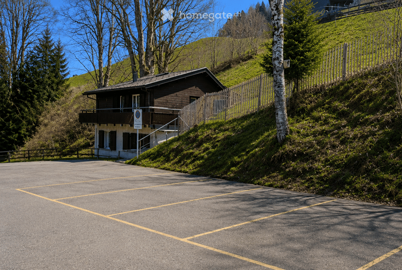A two-story wooden house with a balcony, situated on a hillside with a parking area in front.