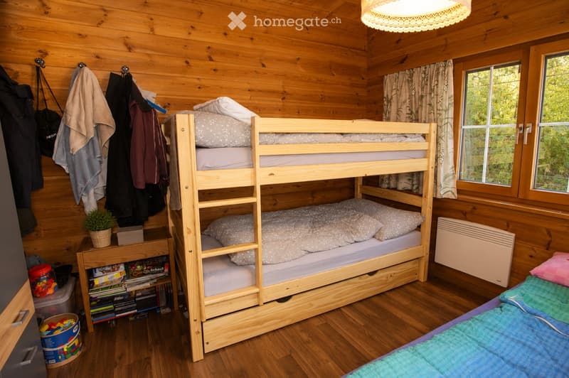 Cozy wooden bedroom with bunk bed, small table with potted plant, storage box, and books. Wooden walls and floor, large windows with curtains, radiator, and hanging clothes.