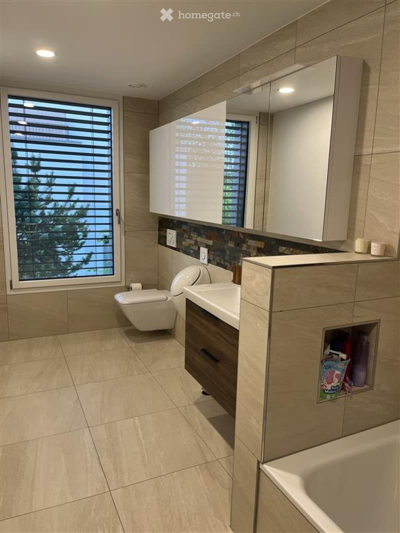 Modern bathroom with beige tiles, a large window with blinds, a toilet, a sink with a wooden cabinet, a mirror, and a bathtub.
