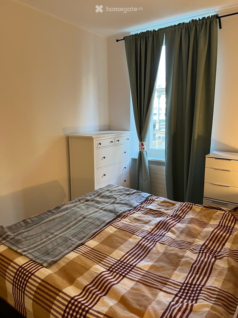 A cozy bedroom with a neatly made bed, a dresser, and a window with curtains.