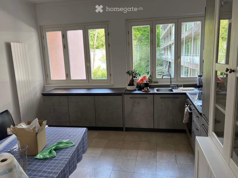 Modern kitchen with grey cabinets, tiled floor, and large windows. Features a sink, faucet, oven, and a small table with a green cloth.