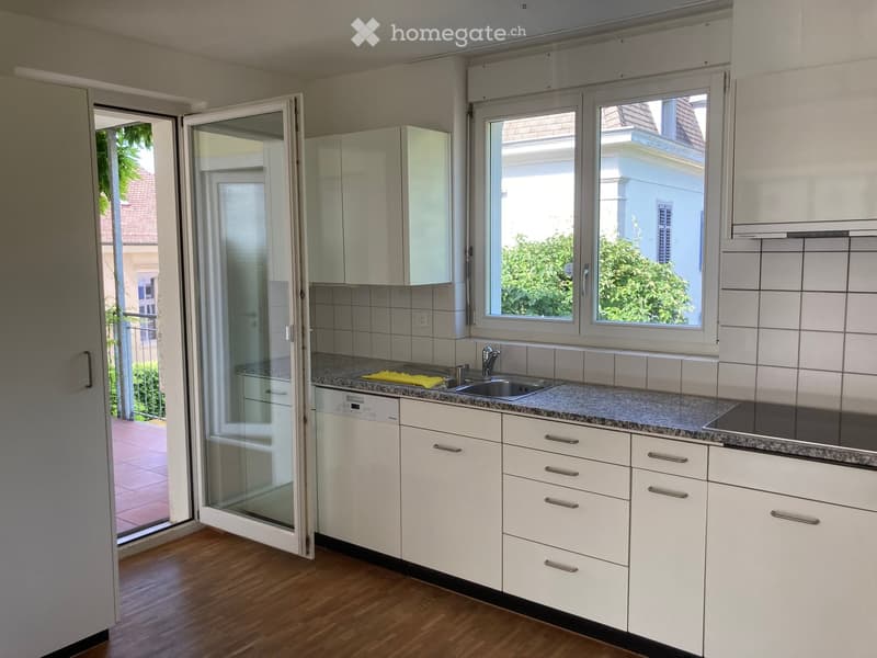 Modern kitchen with white cabinets, granite countertop, stainless steel sink, and dishwasher. Large window and sliding glass door lead to a balcony.