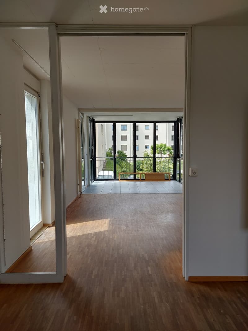 Empty hallway with wooden flooring, white walls, and large windows leading to a balcony area with tiled flooring and a bench.