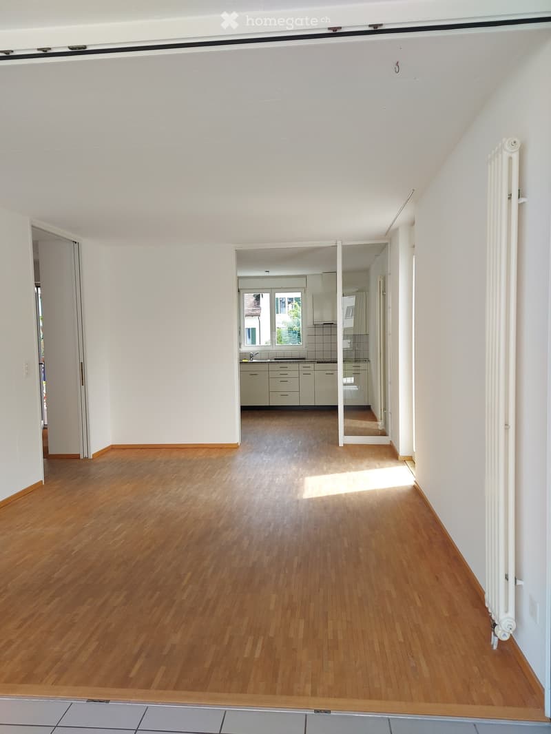 Empty room with wooden flooring and white walls, featuring a radiator on the right side and an open doorway leading to a kitchen area with cabinets and a window.
