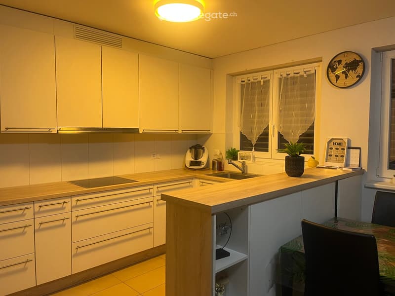 Modern kitchen with light-colored cabinets, a central island, and a sink area. Includes a blender, potted plant, and a clock on the wall.