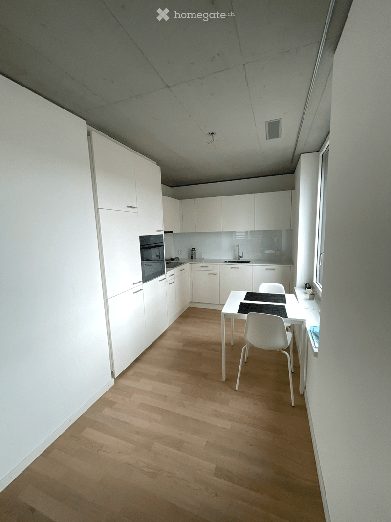 Modern kitchen with white cabinets, wooden flooring, a small dining table with chairs, and built-in appliances including an oven and sink.