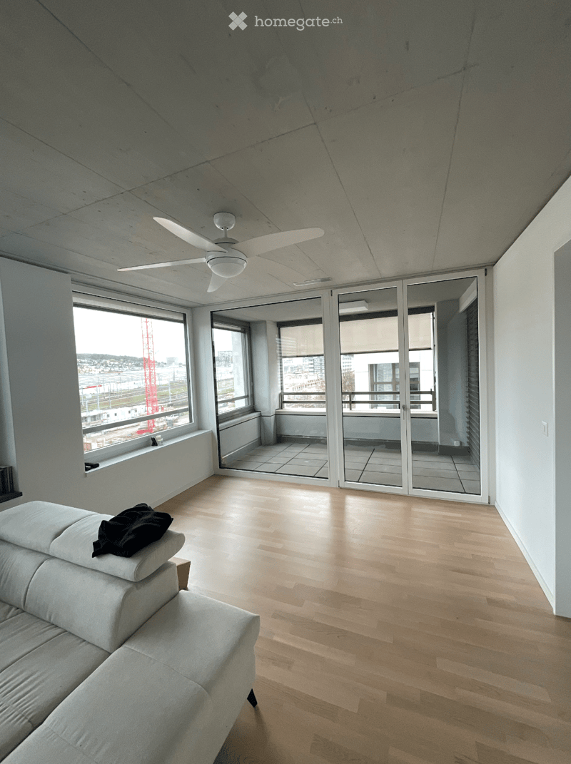 Modern living room with light wooden flooring, large windows, and a sliding glass door leading to a balcony. A grey sectional sofa is positioned near the window, and a ceiling fan is visible.
