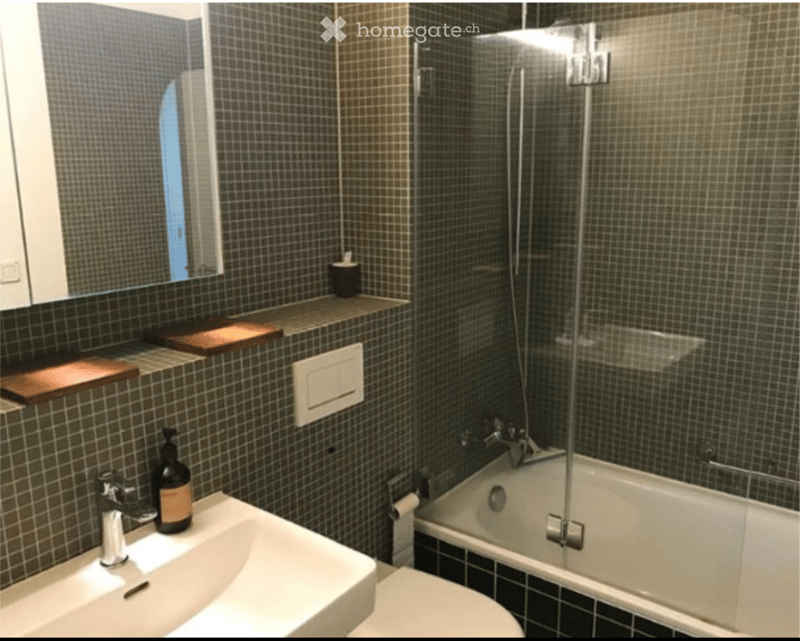 Modern bathroom with dark tiled walls, a white sink, a bathtub with a glass shower enclosure, and a toilet. A mirror is mounted on the wall above the sink, and a small shelf holds a bottle and a wooden tray.