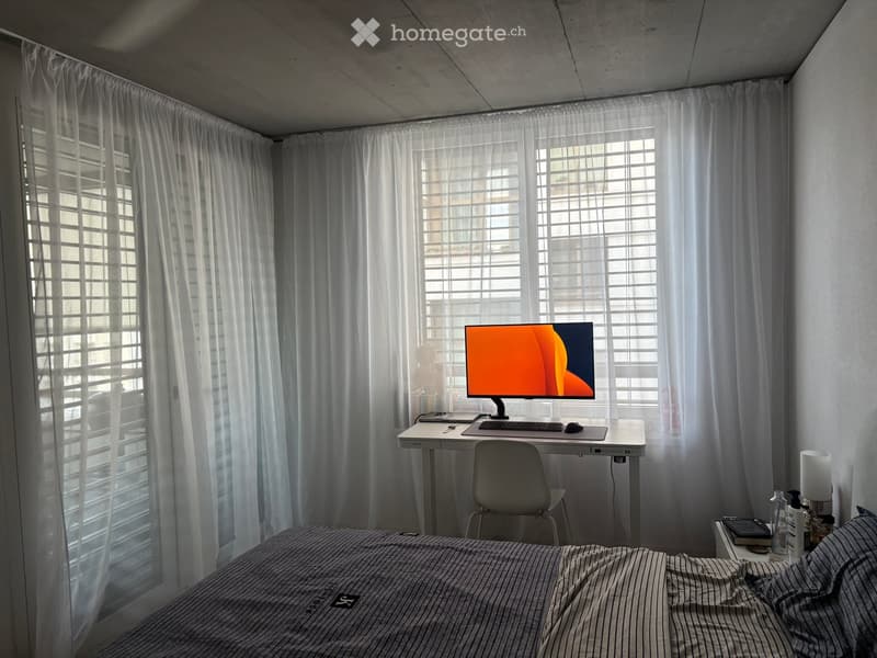 A modern, minimalist bedroom with a bed, desk, and chair. The desk has a computer screen, keyboard, and mouse. A nightstand with a lamp and bottle is visible.