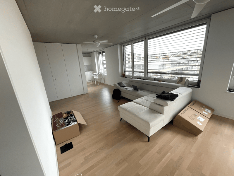 Modern living room with light wooden flooring, white walls, large windows, and a white sectional sofa. Visible items include a laptop, a backpack, and cardboard boxes.