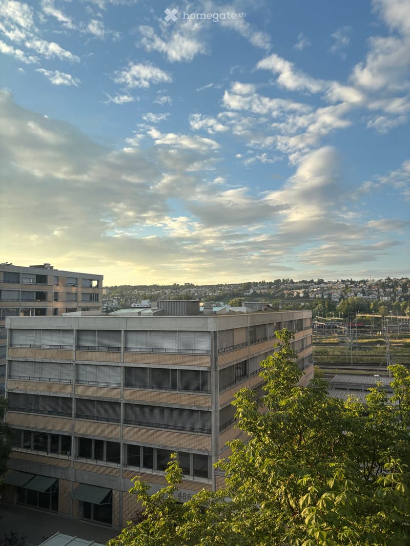 Multi-story building with brick facade, large windows, and a flat roof, surrounded by trees and other structures in an urban setting.