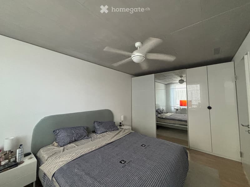 Modern bedroom with a grey upholstered bed, nightstand, mirror wardrobe, and ceiling fan.
