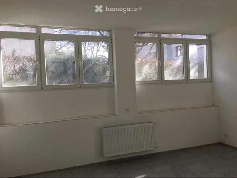 Empty room with bare walls, tiled floor, and large windows allowing natural light. A radiator is mounted on one wall.
