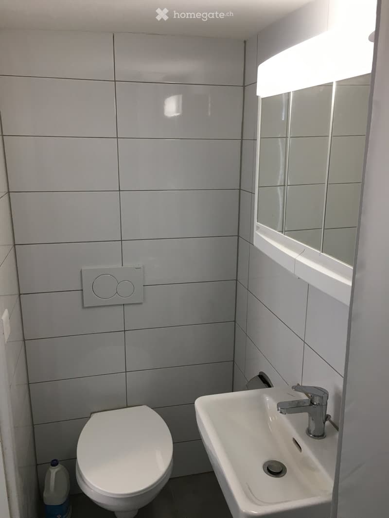 Modern bathroom with white tiled walls, a toilet, a sink with a modern faucet, and a large mirror with built-in lighting above the sink.