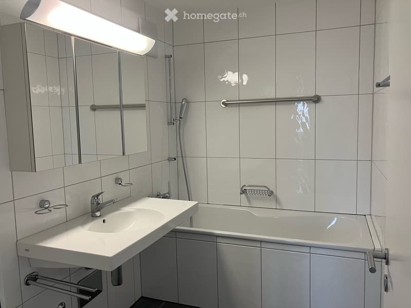 Modern bathroom with white tiles, a sink, bathtub, mirror, and handrails for accessibility.