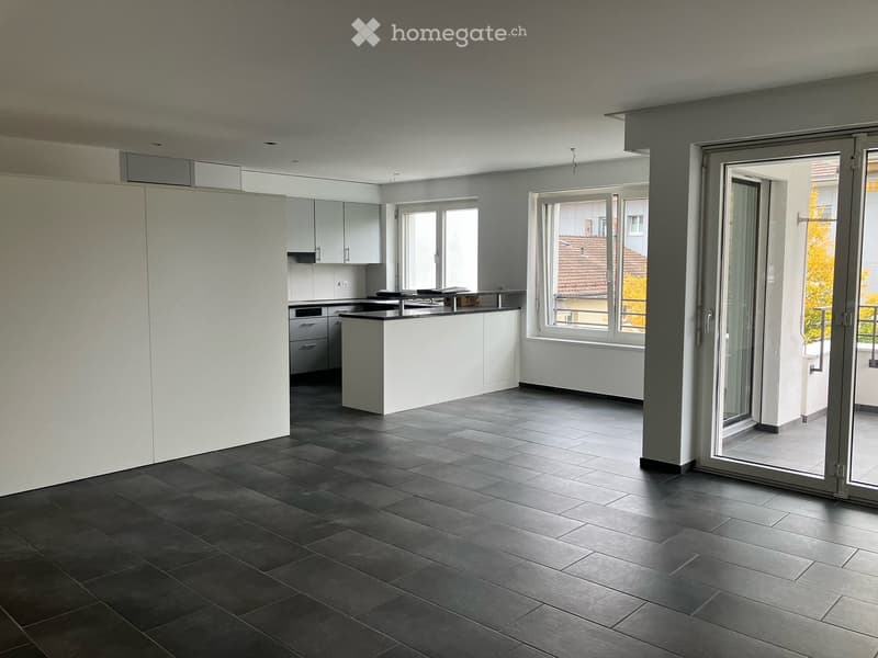 Modern, unfurnished living space with dark tiled flooring, white walls, large windows, and a sliding glass door leading to a balcony. The kitchen area features white cabinets and a countertop.