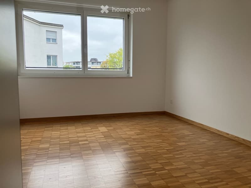 Empty room with wooden flooring and large windows offering a view of neighboring buildings and trees.