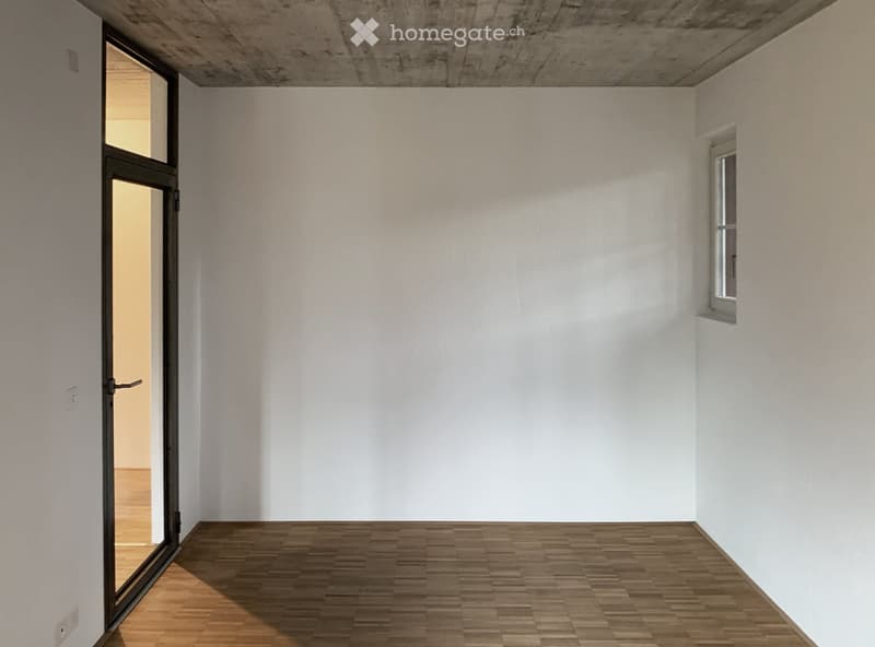 Empty room with white walls, wooden floor, and a large window on one side. A door leads to another room.