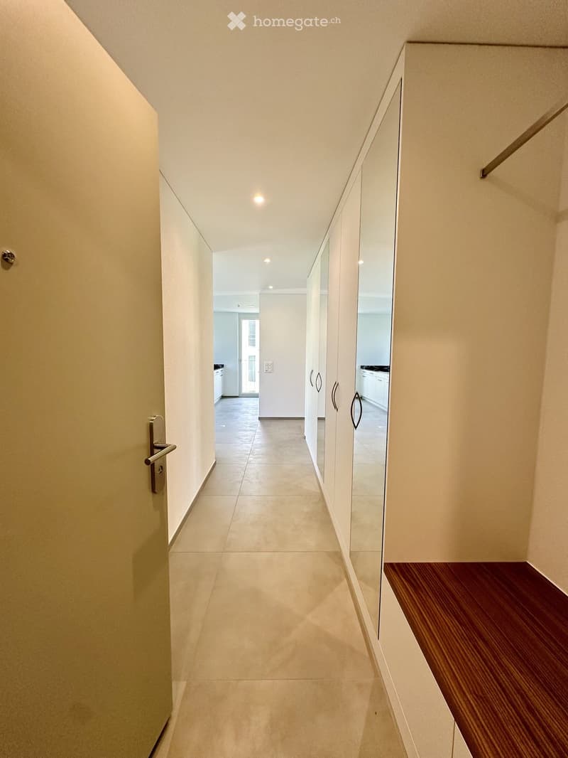Clean, modern hallway with beige tiled flooring, white walls, and built-in mirrored cabinets on one side. A wooden bench is visible on the right side.