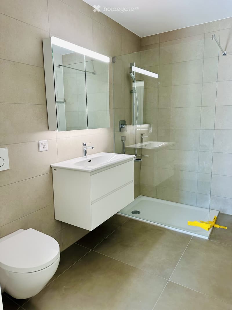 Modern bathroom with beige tiles, a white sink with a cabinet, a toilet, a large mirror with built-in lighting, and a glass-enclosed shower with a modern faucet.