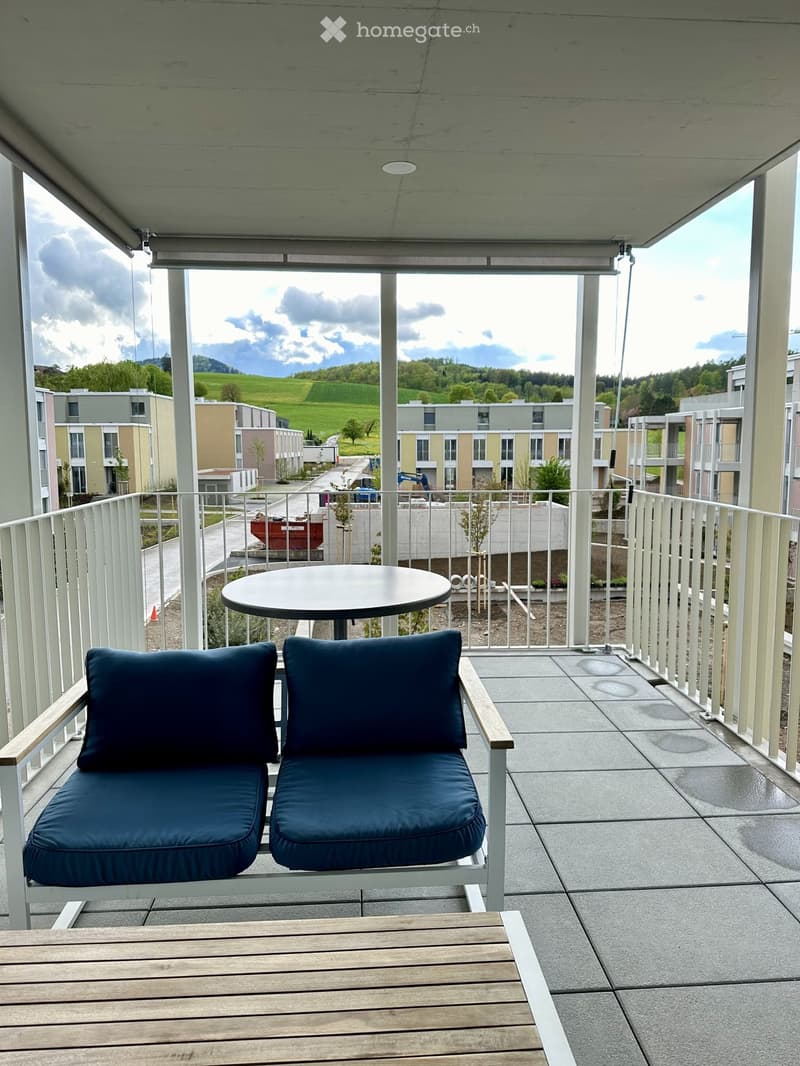 Balcony with a round table, two blue cushioned chairs, and a wooden bench, overlooking a green landscape with buildings and construction.