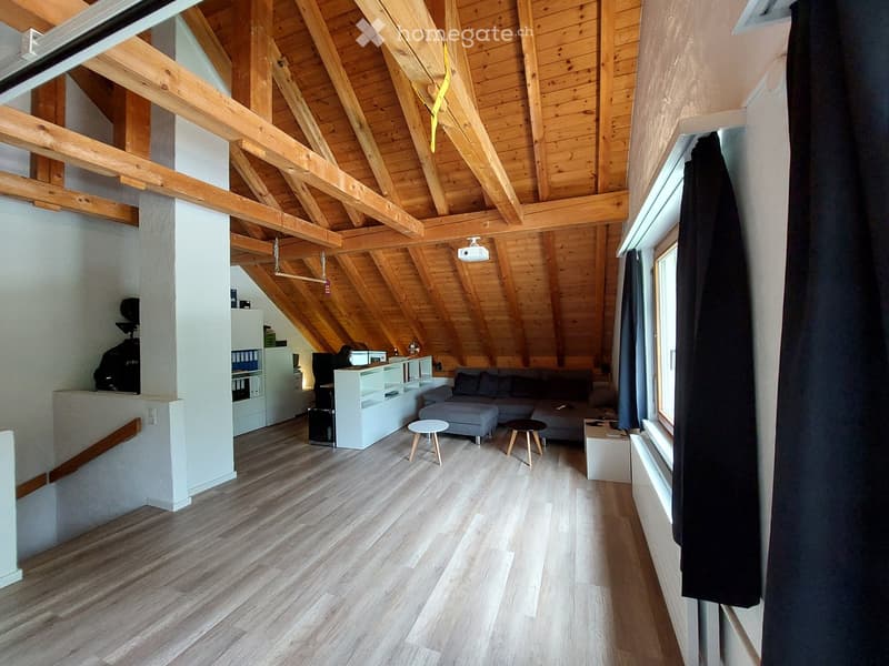 Bright, wooden-beamed attic living space with a grey sectional sofa, two small wooden stools, a white cabinet, and a small table.